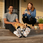 A corgi resting between a man's feet in custom white dog socks, a woman photographing them on a sunny porch